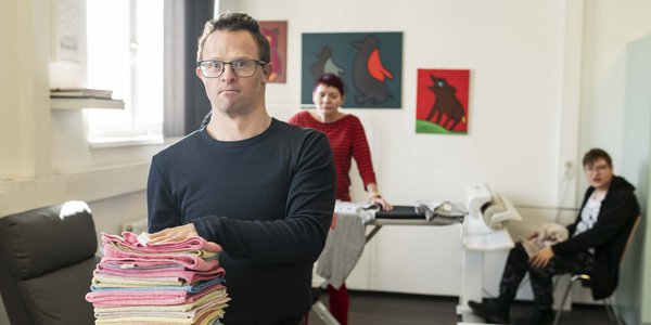 Mensch mit Behinderung mit Wäschestapel im Vordergrund, dahinter zwei weitere Personen an einem Bügelbrett