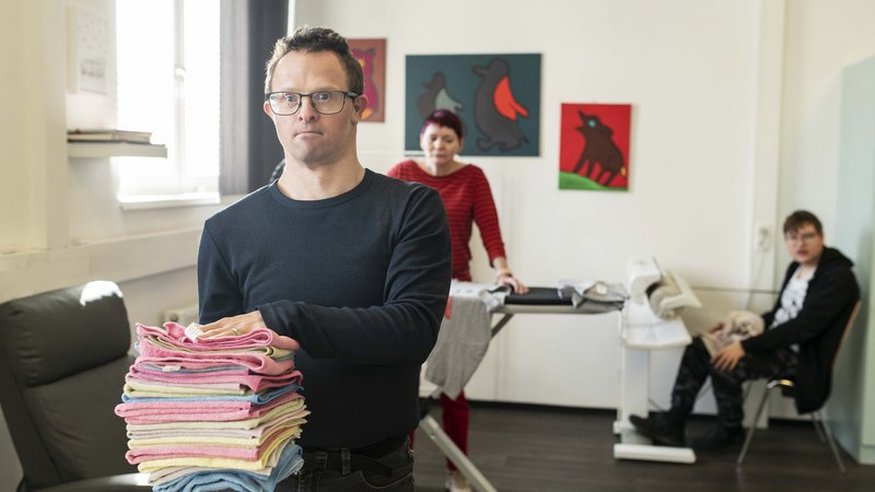 Mensch mit Behinderung mit Wäschestapel im Vordergrund, dahinter zwei weitere Personen an einem Bügelbrett