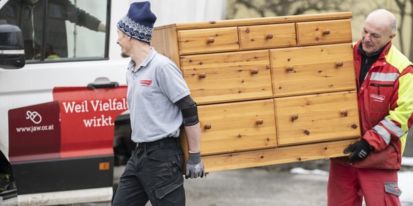 Zwei Männer tragen eine hölzerne Kommode in Richtung eines Vans mit der Aufschrift "Vielfalt wirkt"