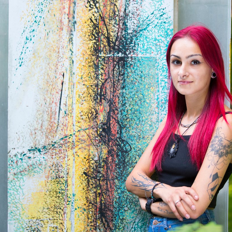 Eine junge Frau mit rosa Haaren und Tattoos vor einer bunten Wand