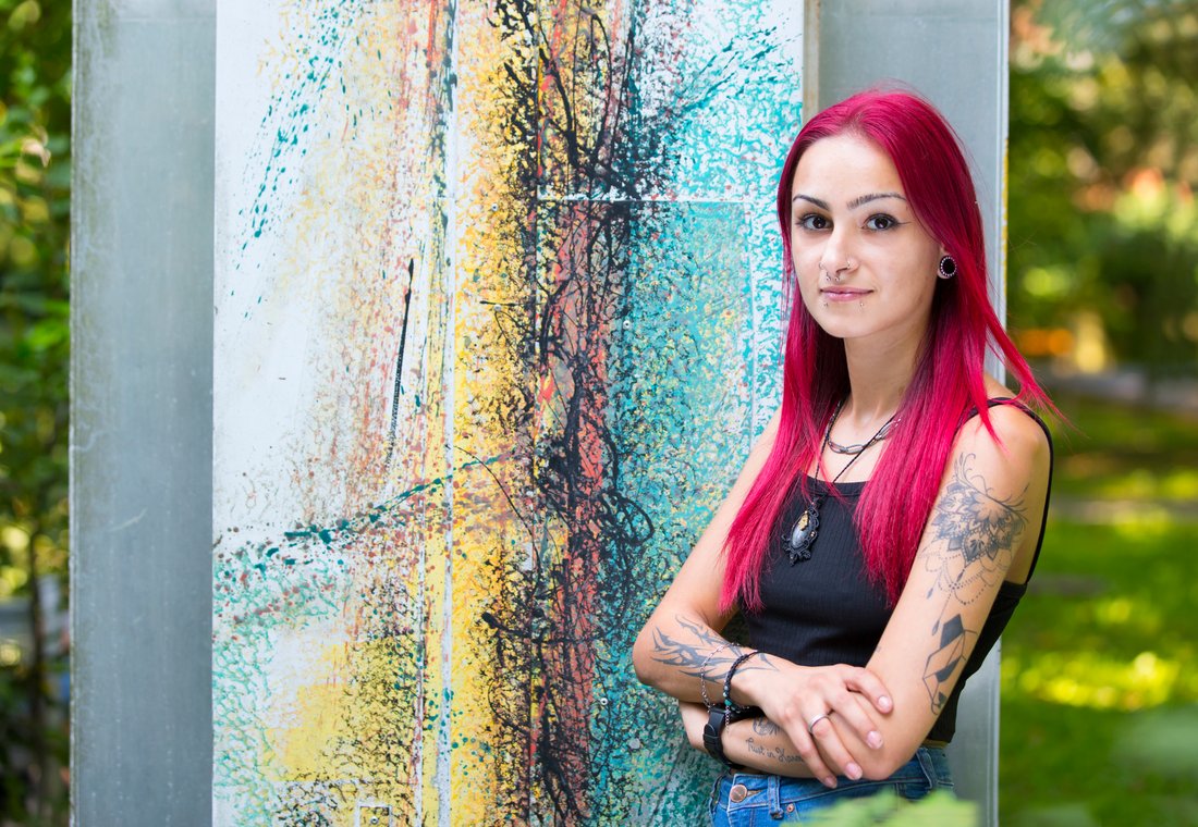 Eine junge Frau mit rosa Haaren und Tattoos vor einer bunten Wand