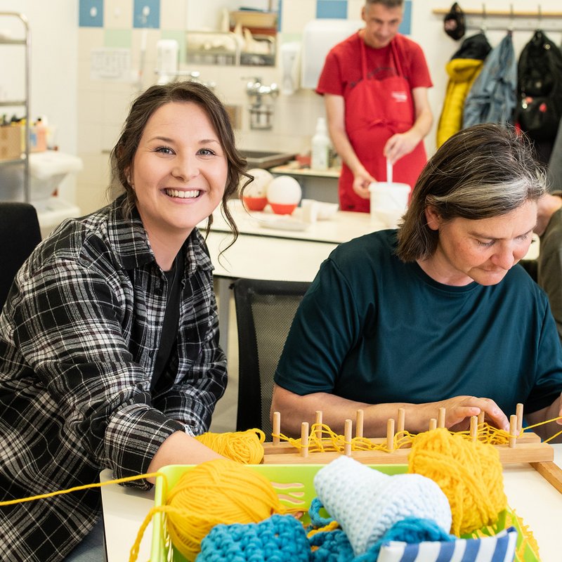 Zwei Frauen stricken mithilfe eines Holzstückes.