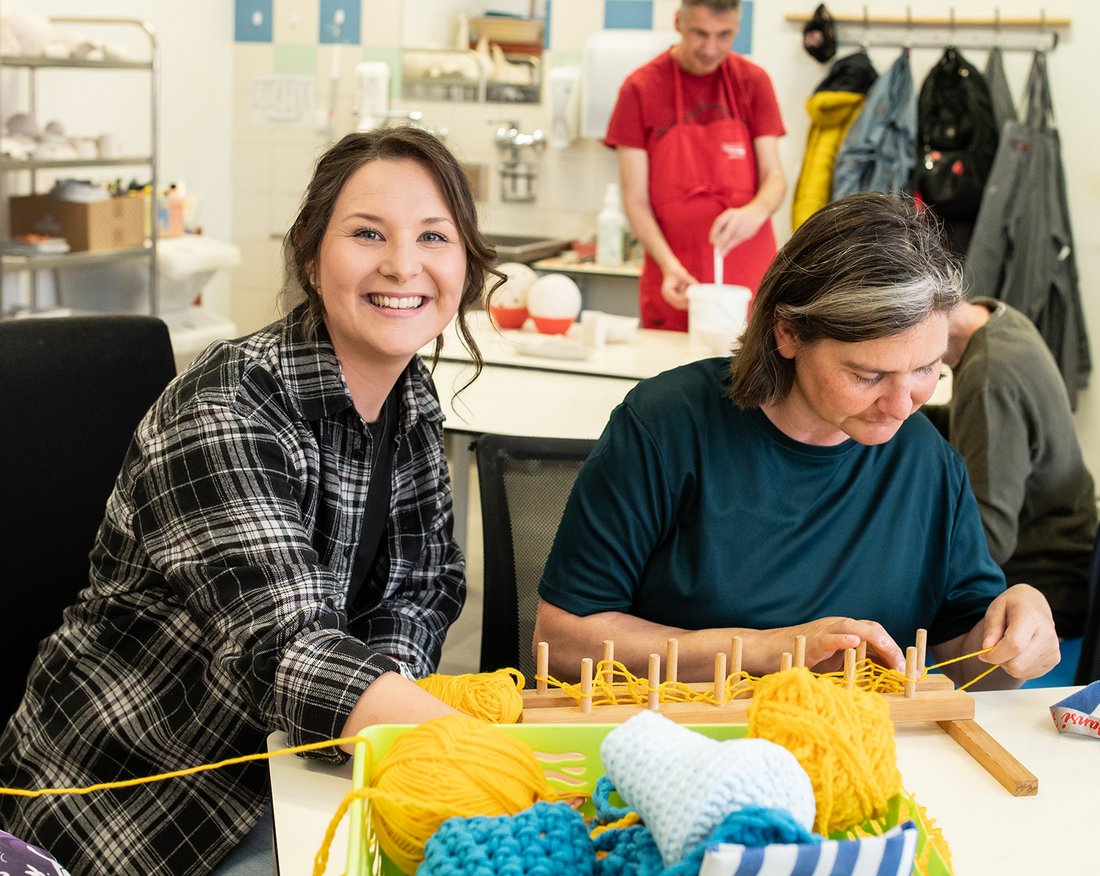 Zwei Frauen stricken mithilfe eines Holzstückes.