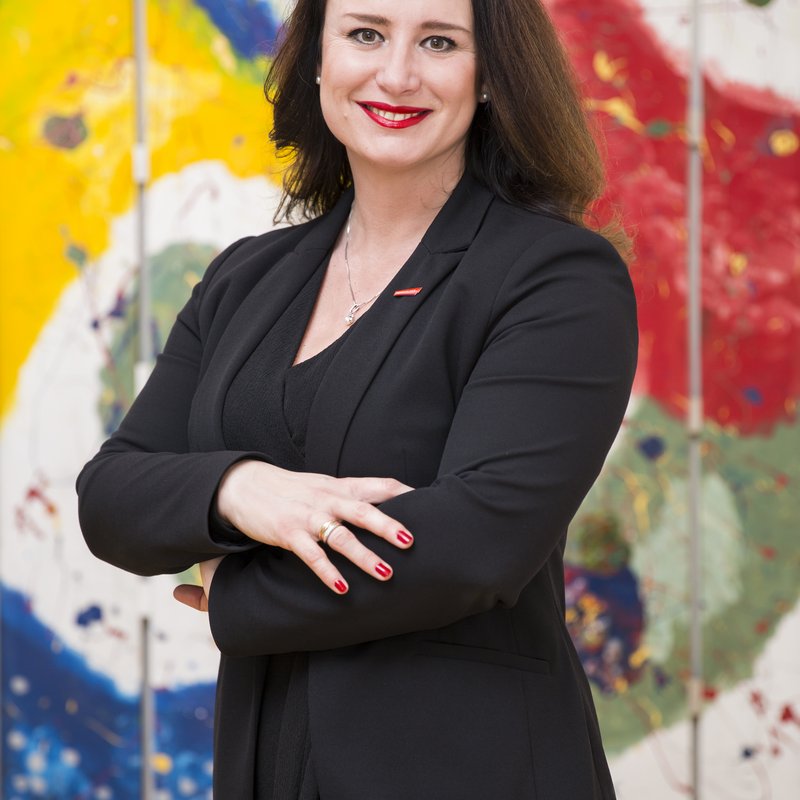 Eine Frau in einem Blazer, die mit verschränkten Armen breit lächelnd vor einem bunten Kunstwerk steht