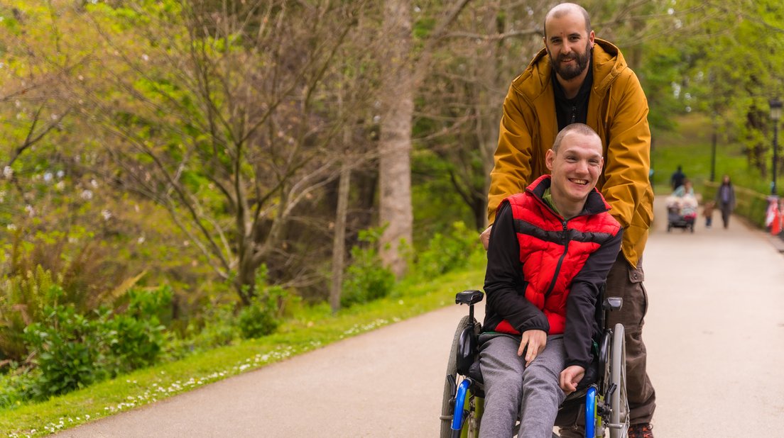 ein Betreuer schiebt einen Mann mit Behinderung im Rollstuhl an, sie freuen sich beide