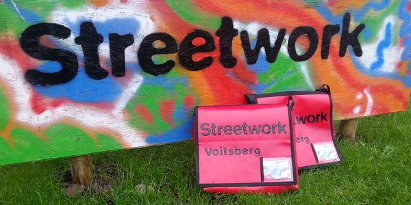 Gras, Wand-Graffiti mit dem Schriftzug "Streetwork", zwei rote Streetwork-Taschen