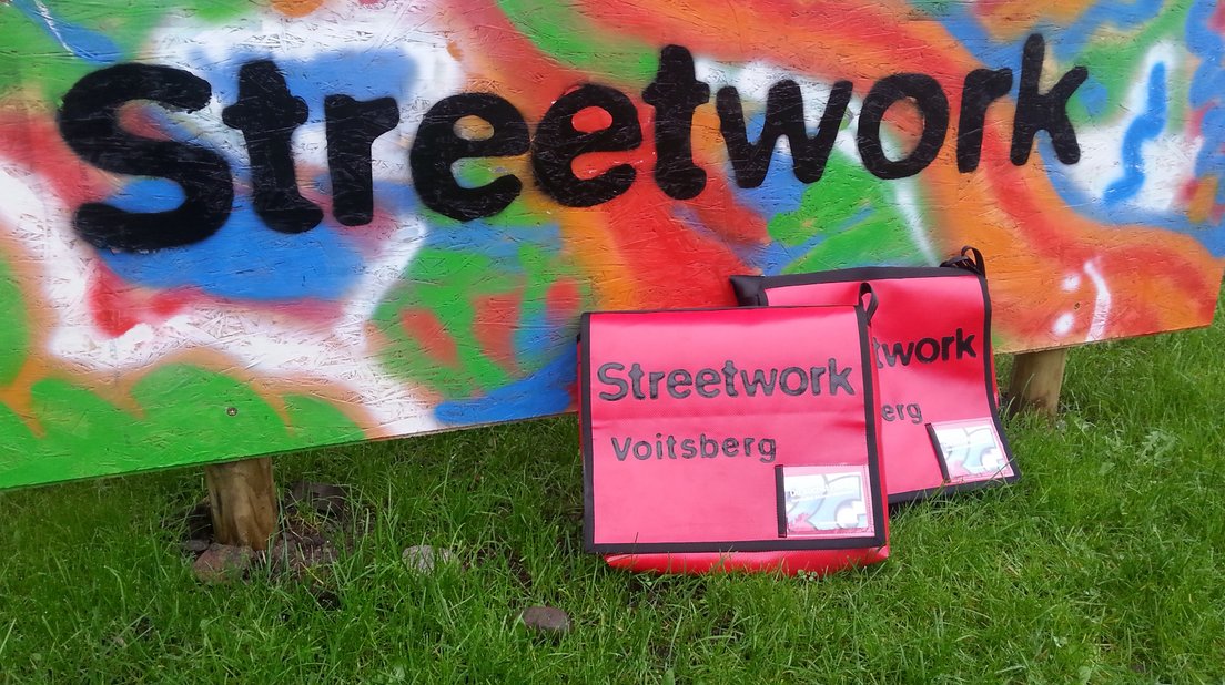 Gras, Wand-Graffiti mit dem Schriftzug "Streetwork", zwei rote Streetwork-Taschen