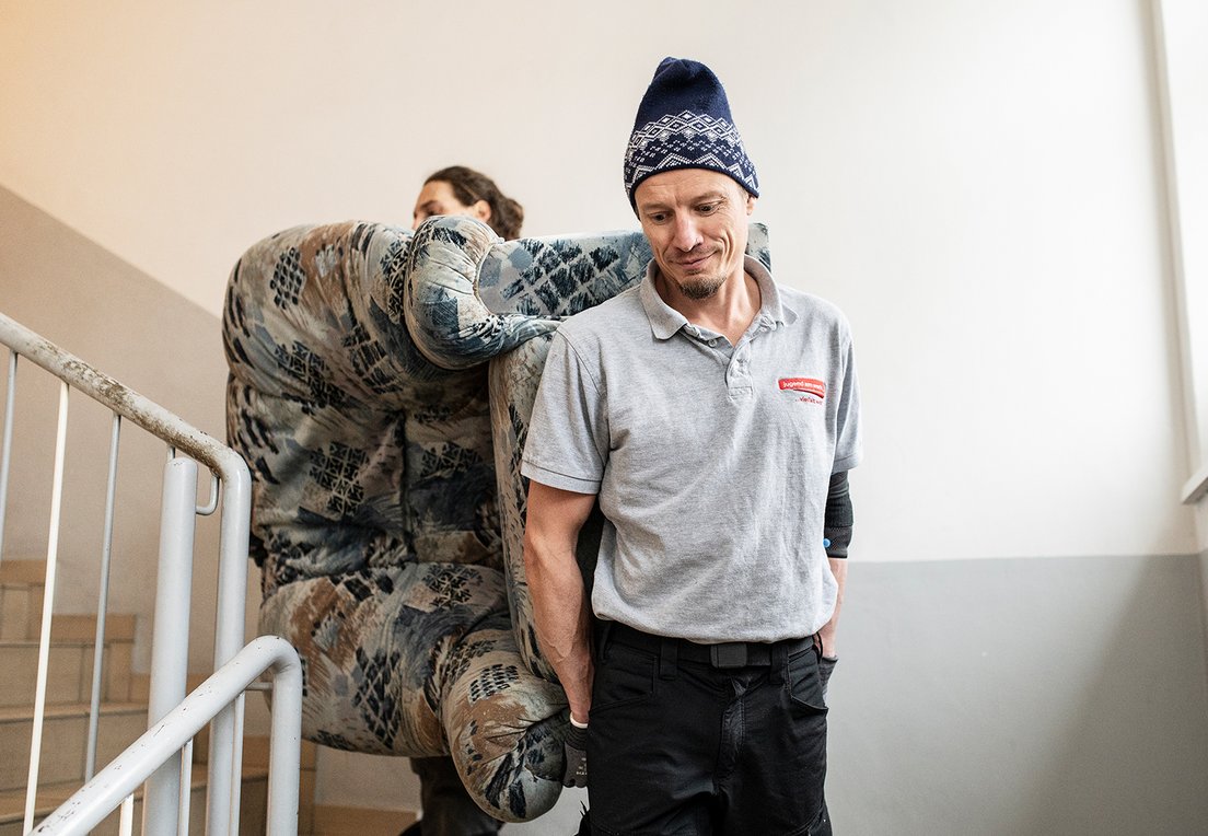 Zwei Männer tragen einen Ohrensessel durch ein Stiegenhaus