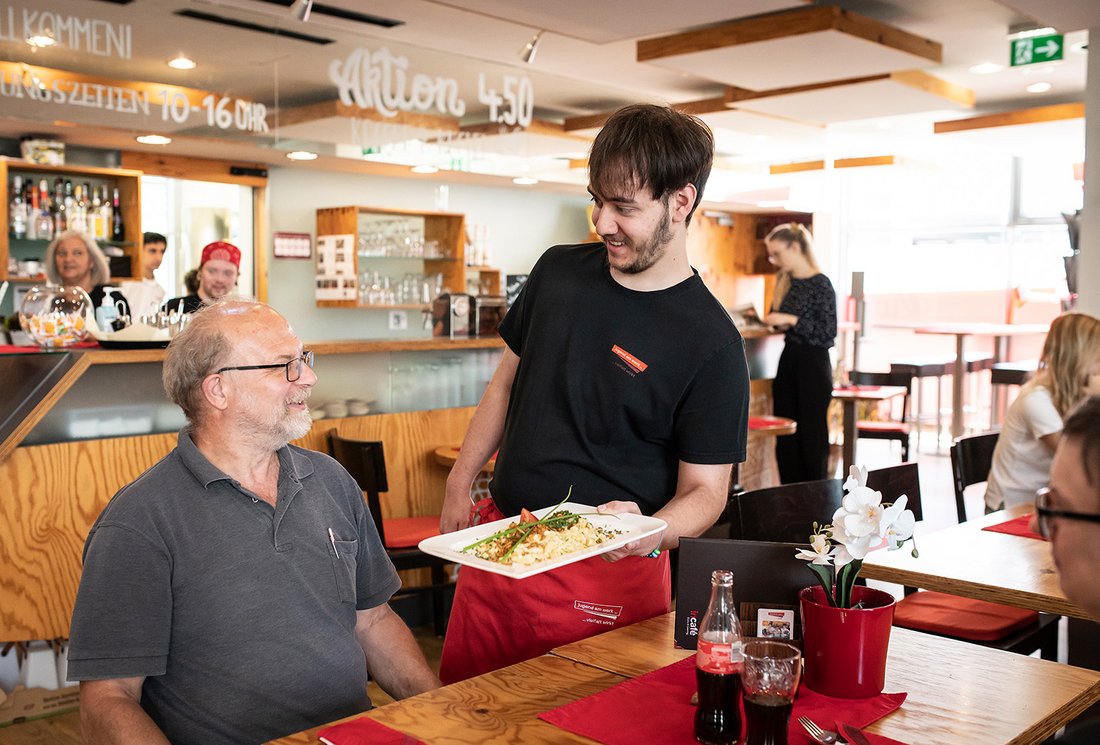 Ein Mann serviert einem Gast ein Mittagsmenü im incafé bei Jugend am Werk Steiermark 