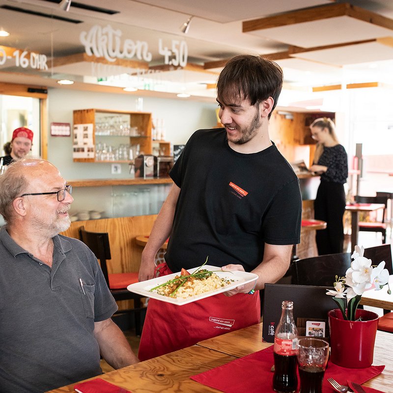 Ein Mann serviert einem Gast ein Mittagsmenü im incafé bei Jugend am Werk Steiermark 