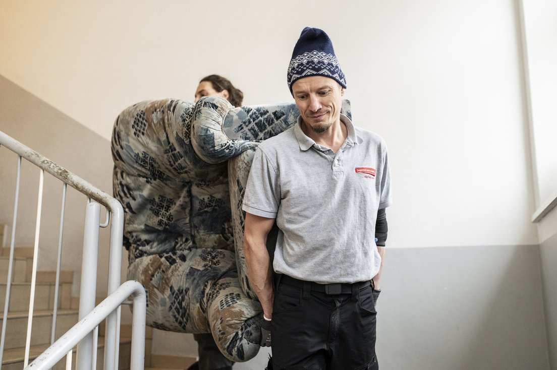 Zwei Männer tragen einen Ohrensessel durch ein Stiegenhaus
