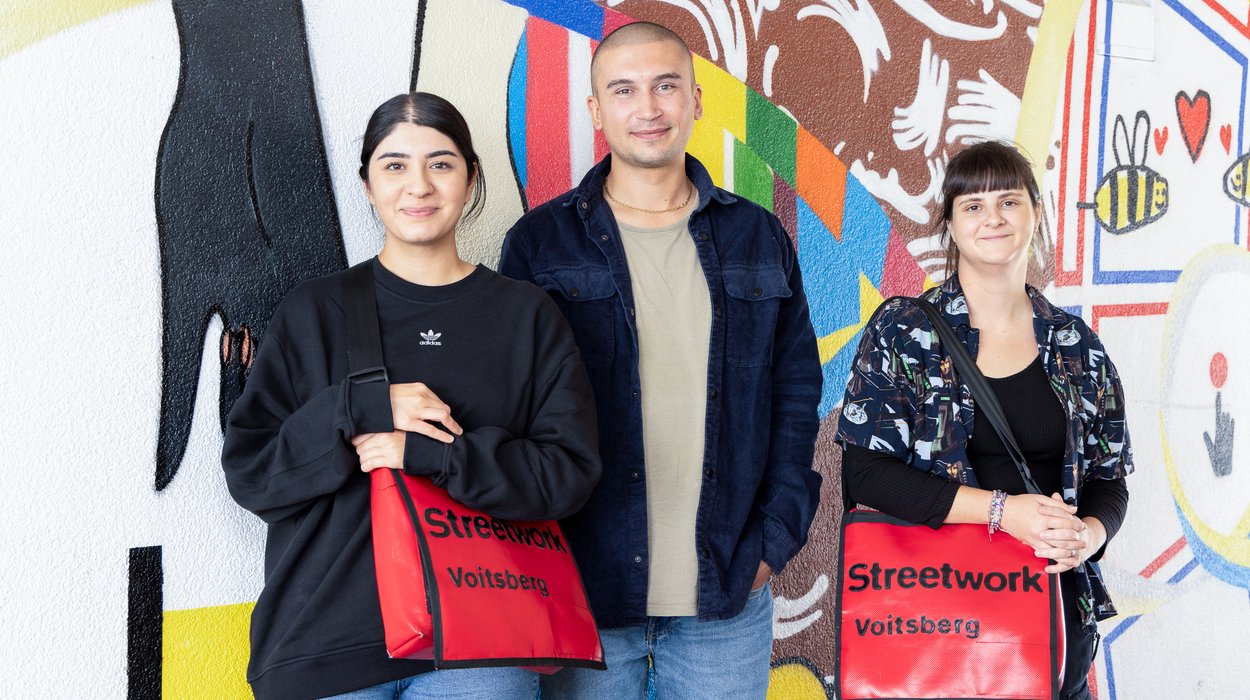 Drei Mitarbeitende von Jugend am Werk vor einer bunten Wand, sie haben Taschen mit der Aufschrift "Streetwork Voitsberg" umgehängt