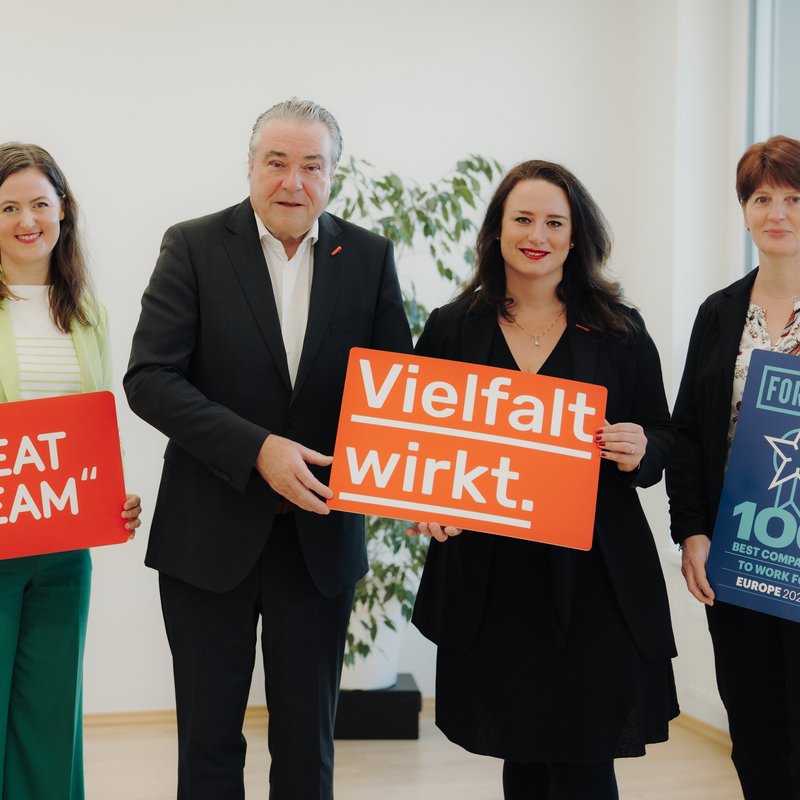 Drei Frauen und ein Mann in Anzug und Blazer halten Schilder mit der Aufschrift "Fortune", "Great Team" und "Vielfalt wirkt" hoch