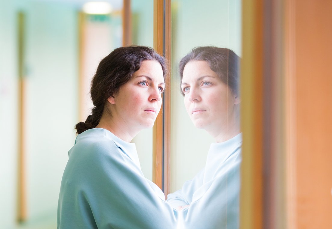 Eine Frau steht vor einem Fenster und sieht hinaus, ihr eigenes Gesicht ist in der Spiegelung zu sehen