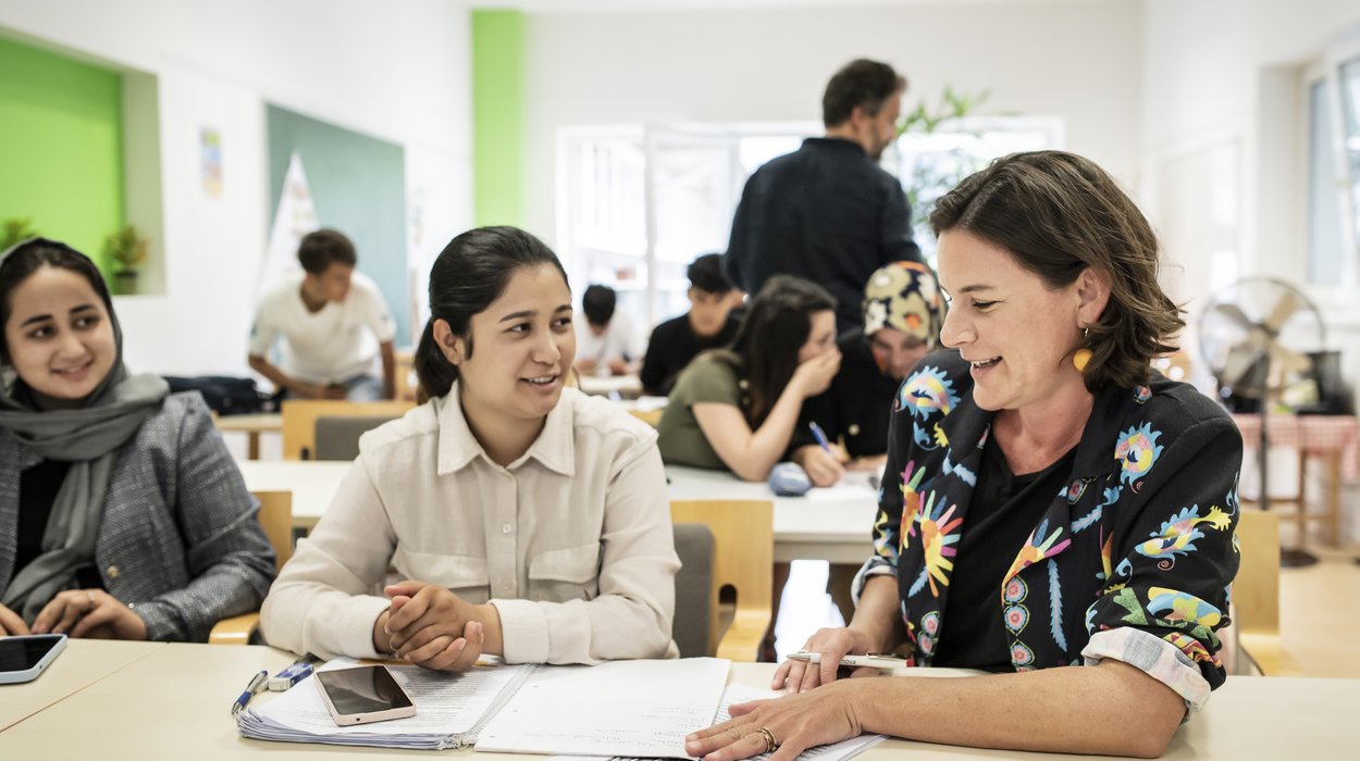 Zwei Frauen an einem Tisch in einem vollen Klassenzimmer. Eine unterrichtet die andere.