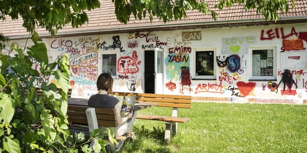 ein Mädchen liest ein Magazin vor einem Haus, das voller Graffiti ist, auf einer Gartenbank mit Tisch unter einem Baum