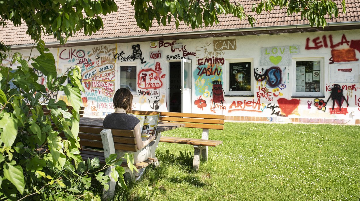 ein Mädchen liest ein Magazin vor einem Haus, das voller Graffiti ist, auf einer Gartenbank mit Tisch unter einem Baum
