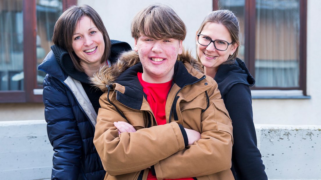 ein Jugendlicher mit Behinderung und zwei Betreuerinnen stehen freudig vor einem Gebäude