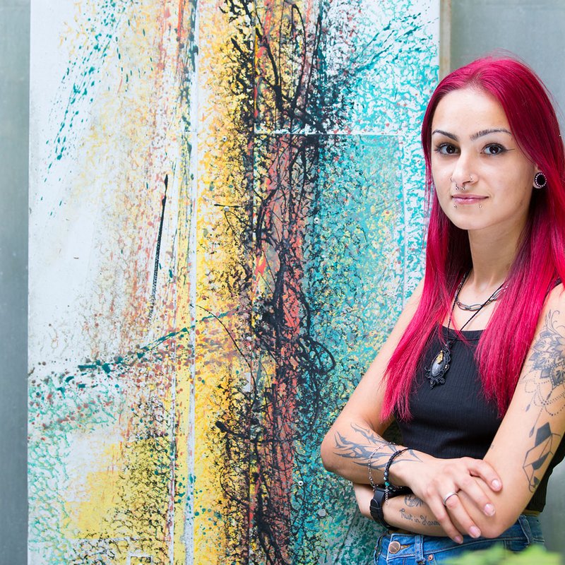 Eine junge Frau mit rosa Haaren und Tattoos vor einer bunten Wand
