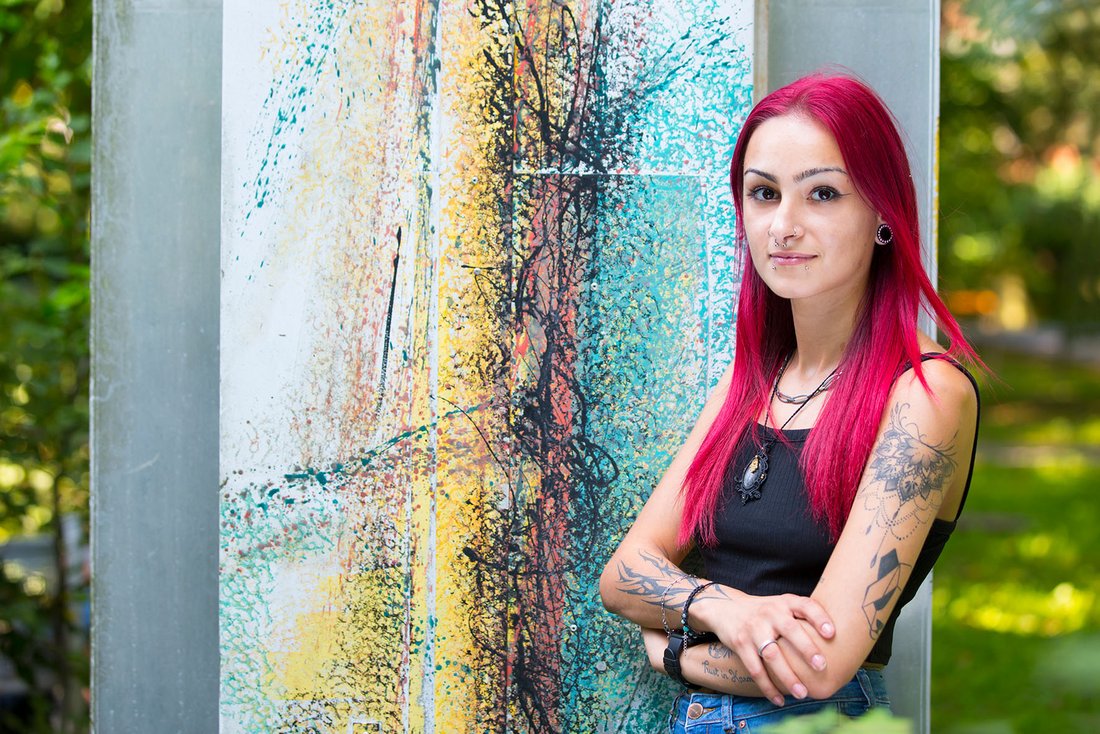 Eine junge Frau mit rosa Haaren und Tattoos vor einer bunten Wand