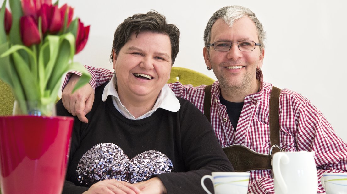 zwei Personen mit Behinderungen sitzen lachend nebeneinander in einem schönen Raum